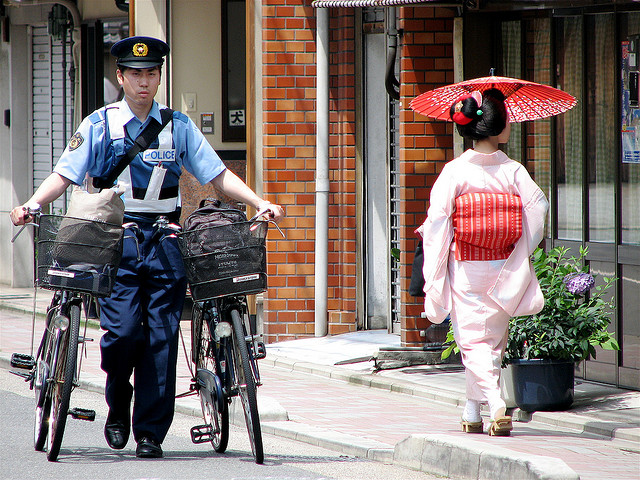 A Japanese policeman pushing two bicycles passes a geisha with a parasol