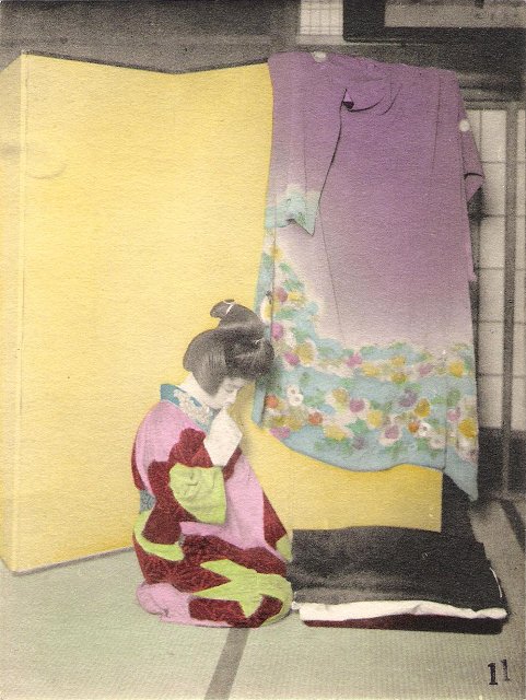 A women in a nightgown with a formal Japanese hairstyle kneeling in front of a screen on which a kimono is hung